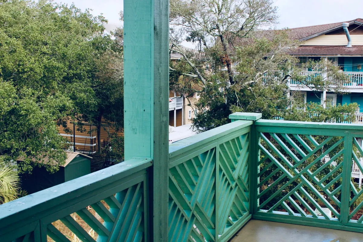 This exterior image captures a balcony with a distinctive green railing overlooking lush surrounding trees and glimpses of neighboring buildings. The scene suggests a tranquil spot where guests can unwind while staying closely connected to the outdoors. Soft natural light filters through the trees, creating a relaxed and inviting atmosphere perfect for enjoying the beachy environment.