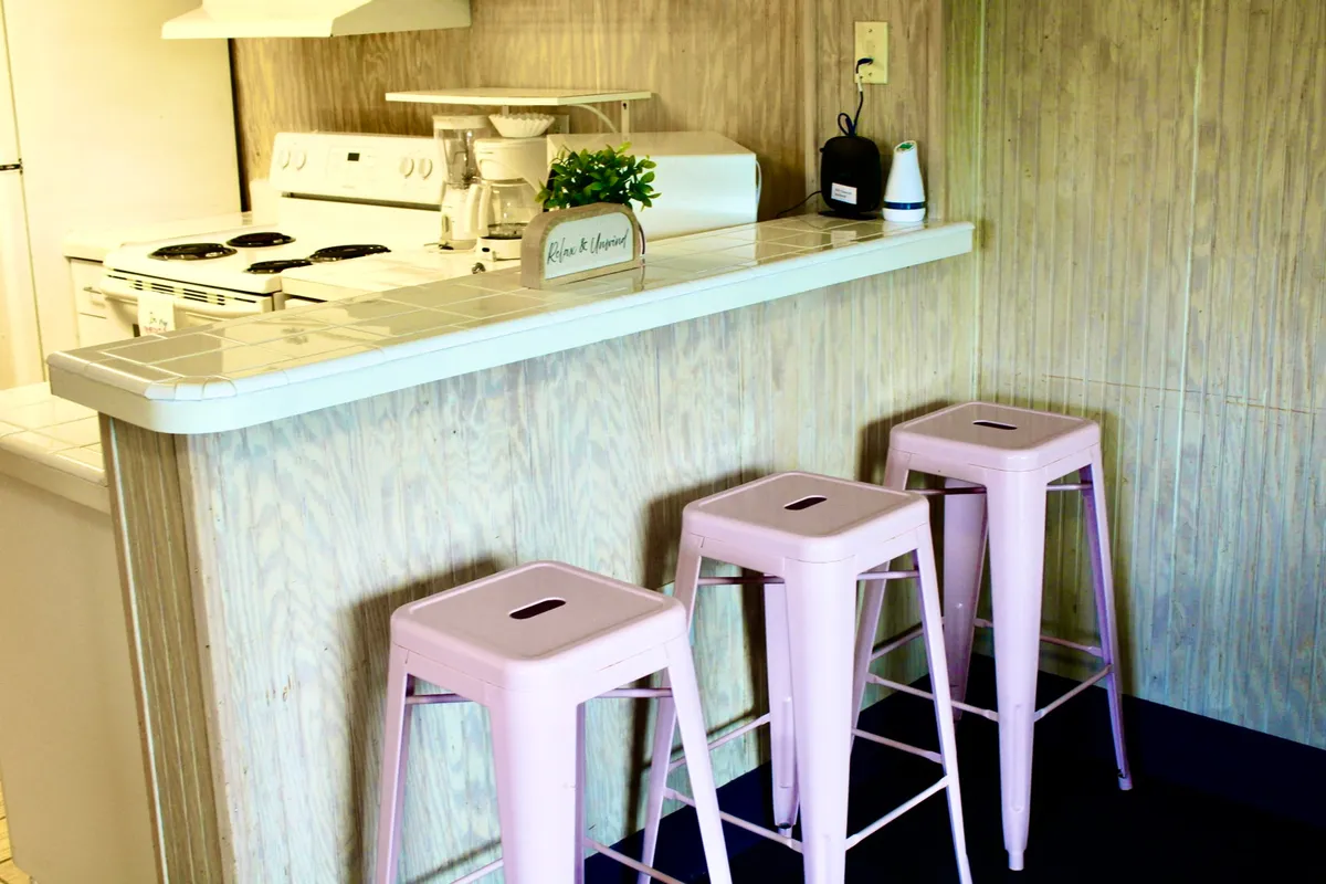 The image showcases the bright and inviting interior of the Ocean Surf Club-B2 studio, with a modern kitchen island at center stage, surrounded by cheerful pink bar stools and a splash of greenery from a decorative plant. Stainless steel appliances and an open layout suggest a space perfect for cooking, dining, and gathering—ideal for both family meals and relaxing getaways.