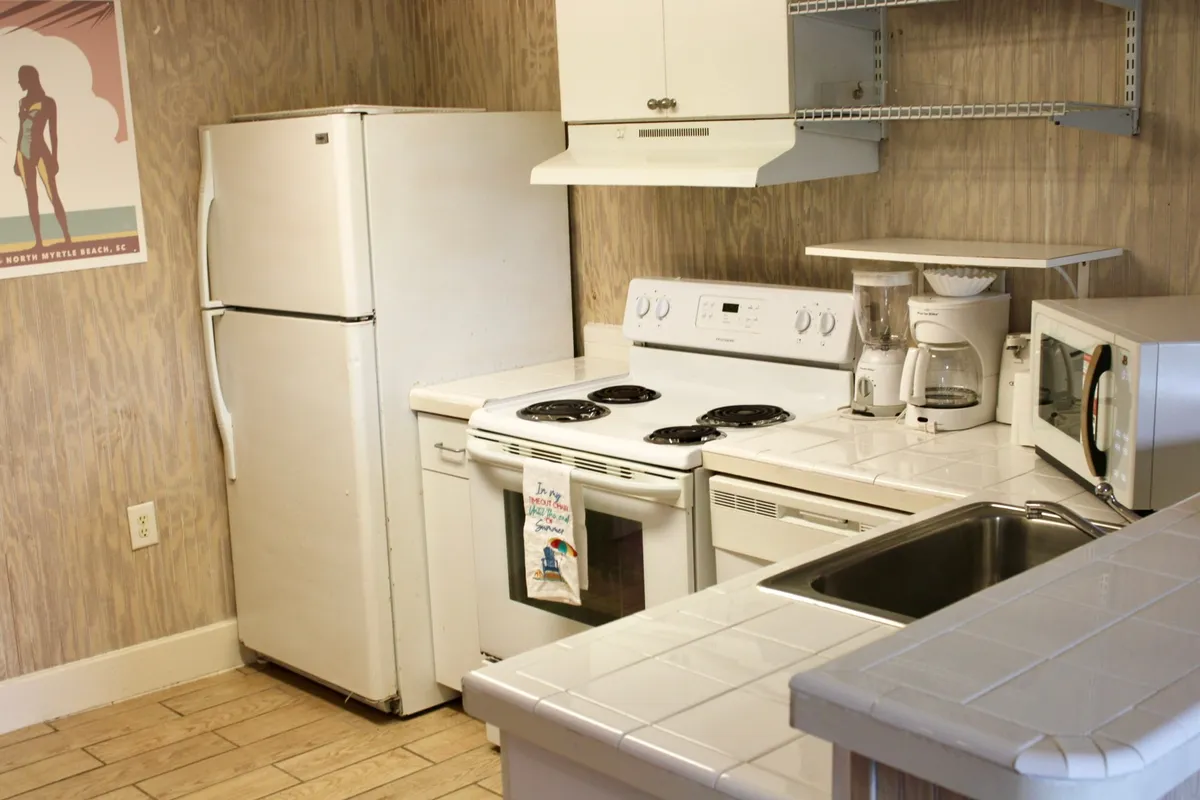 A bright and practical kitchenette featuring white appliances including a refrigerator and oven, tile countertops, a kitchen sink, and a wall poster. The space feels inviting, with clear evidence that it’s set up for cooking, baking, and hosting meals. Utensils and kitchenware are visible, emphasizing functionality.