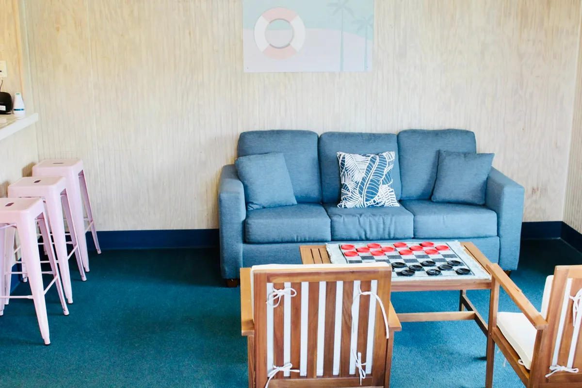 A bright, inviting living area featuring a blue sofa, wooden coffee table with a checkers game set up, and pink bar stools at a breakfast bar in the background. The space is designed for relaxation, socializing, and casual fun, with soft furnishings that add a laid-back, welcoming vibe.