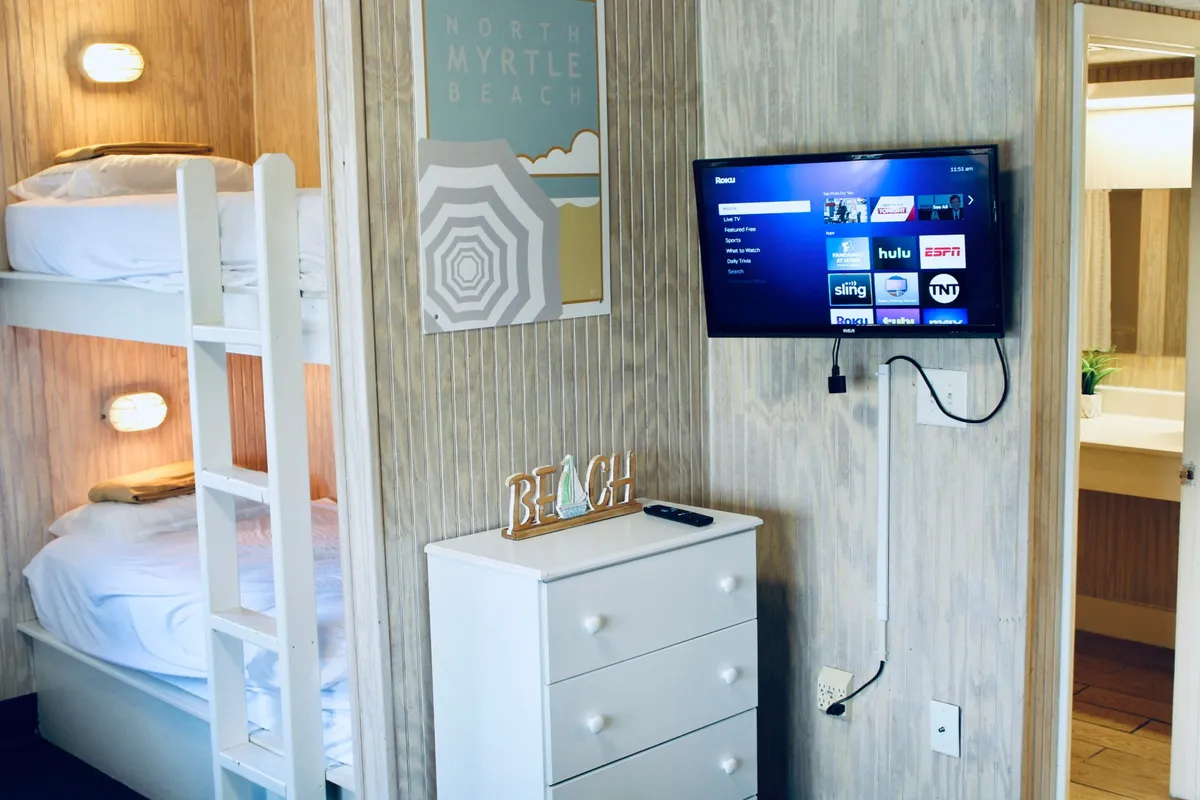 This image centers on a cozy corner of the open-plan living and sleeping area, featuring bunk beds, a television, and charming beach-themed decor such as a 'BEACH' decorative sign and wall art labeled 'NORTH MYRTLE BEACH.' The playful setup suggests space for small families or groups, with comfortable spots for relaxing, watching movies, or chatting together.